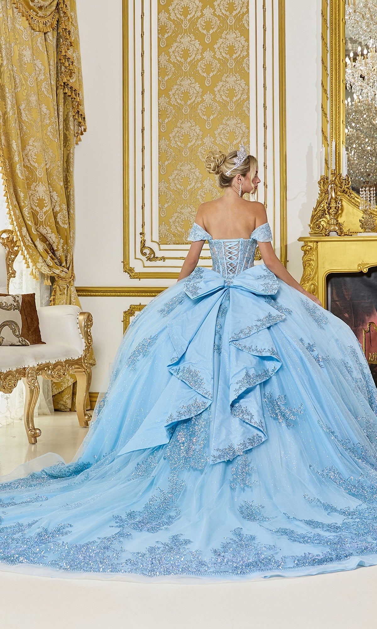 A woman faces away in an opulent, gold-accented room, wearing Quinceanera Dress 5123 By Dancing Queen—an off-the-shoulder light blue gown with a long, ornate train.